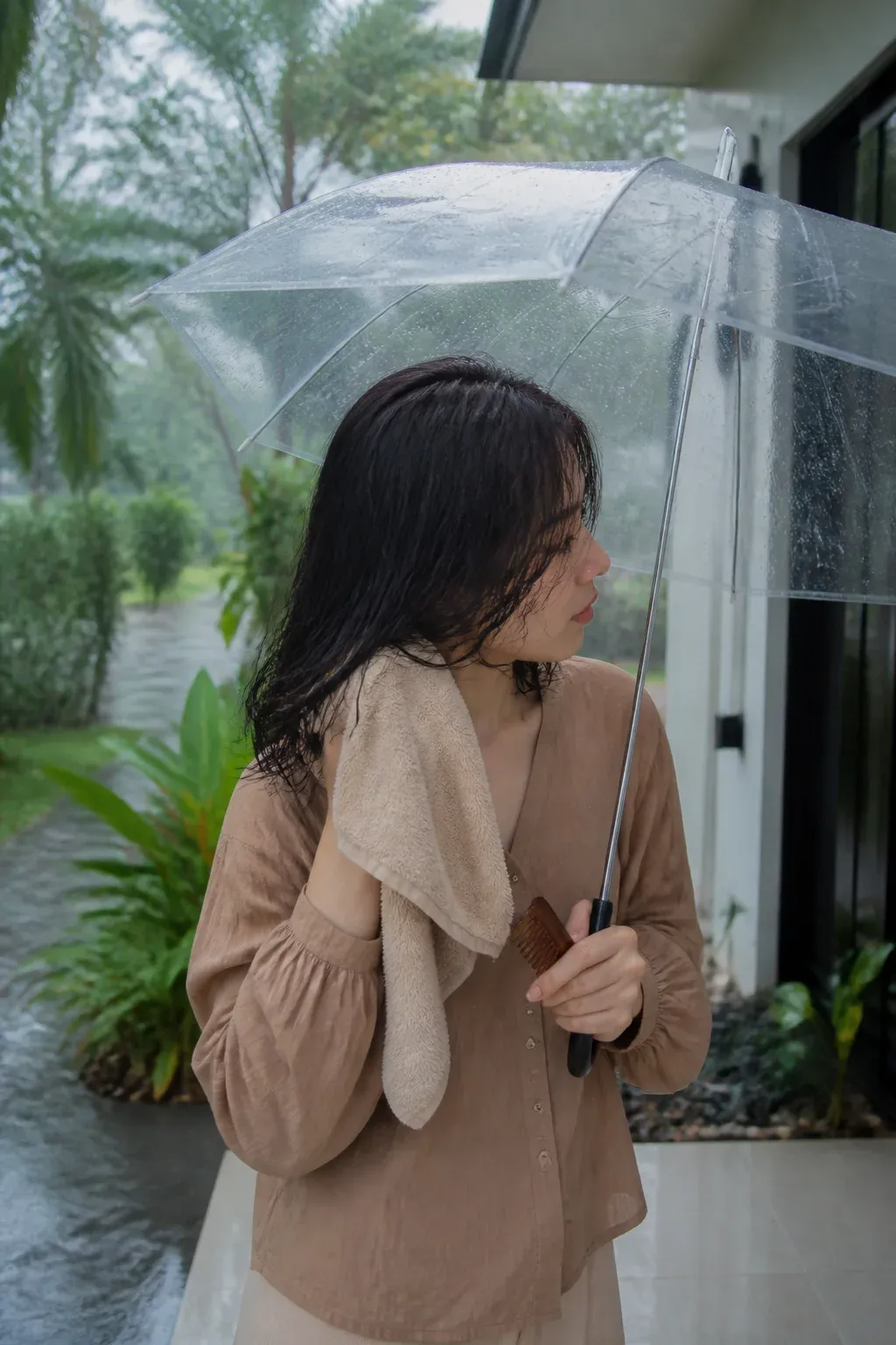 A person under a clear umbrella gently blotting damp dark hair with a towel during rain